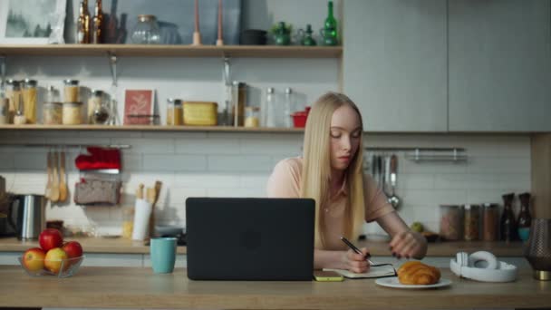 Chica atractiva estudiante que estudia en línea sentado en la mesa de la cocina con el ordenador portátil moderno. Mujer joven enfocada haciendo notas en un cuaderno escuchando lecciones educativas en la computadora. Mujer inteligente aprendiendo en casa. - Metraje, vídeo