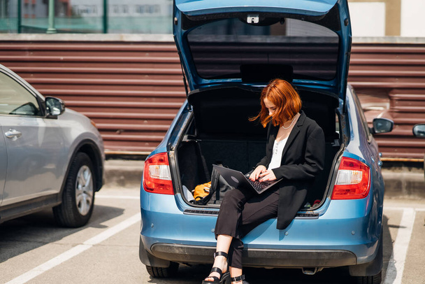 Business attractive young woman working on laptop while sitting in trunk of car while traveling - Valokuva, kuva