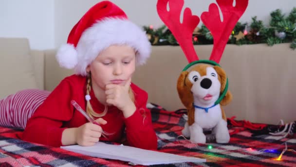 Счастливая маленькая девочка в шляпе Санта-Клауса лежит дома на кровати и пишет письма мечты о подарках Санта-Клаусу. Рождественский список чудес. Ребёнок с рождественскими огнями - Кадры, видео