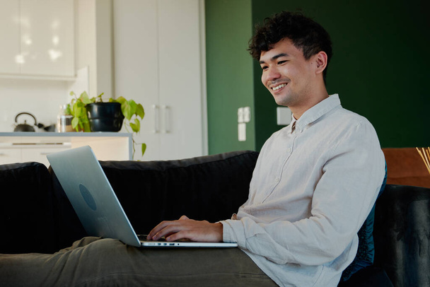 Feliz joven multirracial hombre en camisa usando el ordenador portátil mientras está sentado con los pies en el sofá en la sala de estar en casa - Foto, Imagen