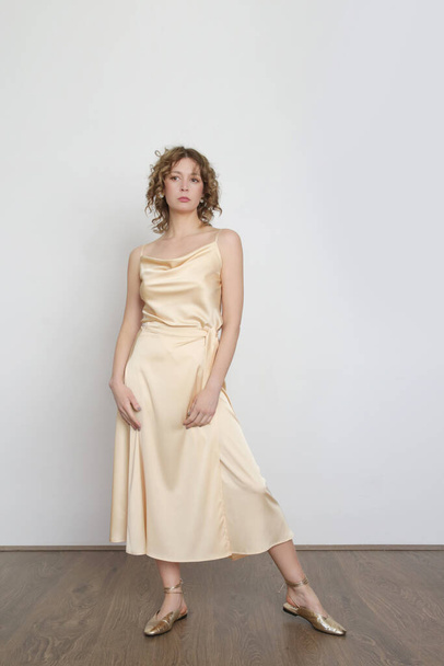 Γυναικείο μοντέλο με χρυσή καμισόλα μεταξωτή μπλούζα και τυλιγμένη φούστα midi. Κομψό μονόχρωμο καλοκαιρινό ντύσιμο. Φωτογραφία στούντιο μόδας. - Φωτογραφία, εικόνα