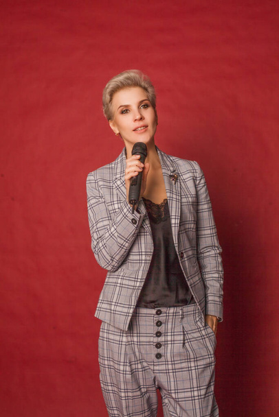Attractive happy glamorous blonde short haired host singer blogger woman in checked suit and white hills holding microphone in hand on red wall background - Fotografie, Obrázek