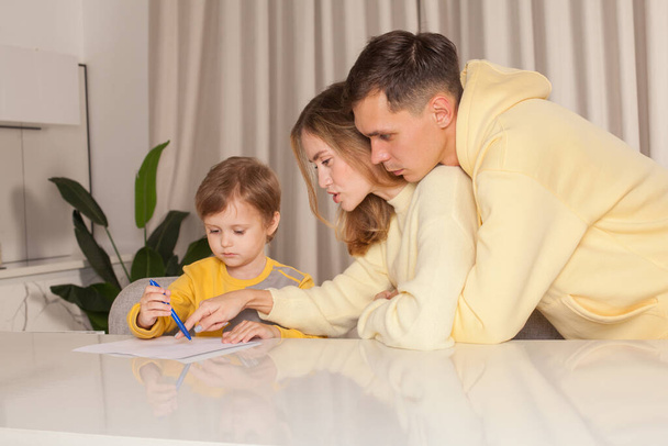 テーブルに座って絵を描く子供の息子と黄色の服の母と父 - 写真・画像