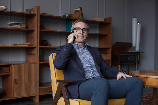 Sério preocupado idoso empresário em terno azul e óculos falando em seu telefone celular no fundo da parede cinza  - Foto, Imagem