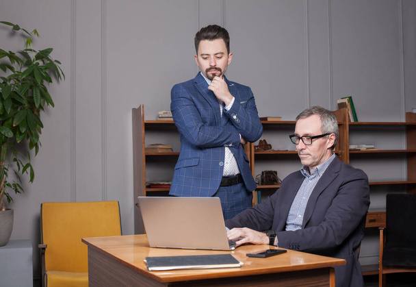 Зрелый седой бизнесмен в синем костюме и очках с коллегой с пенсией думает о новой работе ноутбука в офисе - Фото, изображение