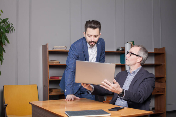 Зрелый бизнесмен в синем костюме показывает свой бизнес-план на ноутбуке своему молодому коллеге в офисе - Фото, изображение