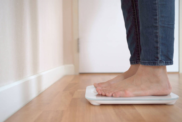 Unrecognizable woman stepping on the digital display weighting scale to measure her body weight and overall body mass index. Health care and wellbeing concept in modern people. - Фото, изображение