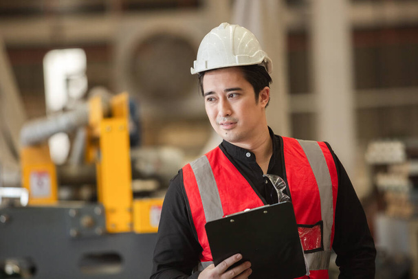 Portret młodego inżyniera trzymającego schowek nosi biały kask w mundurze stojącym w przestrzeni przemysłowej. mężczyzna pracownik duża fabryka przemysłowa - Zdjęcie, obraz
