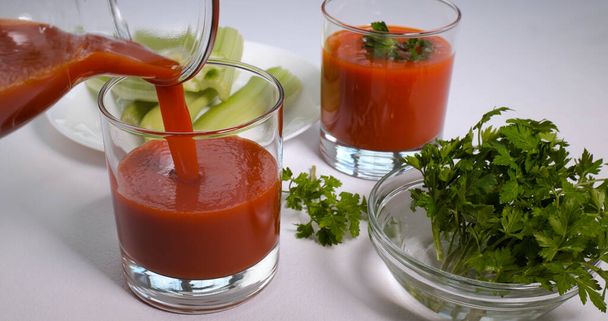 Verser du jus de tomate fraîchement pressé biologique dans un verre. Céleri et persil sur une table blanche. Concept alimentaire sain et végétarien - Photo, image