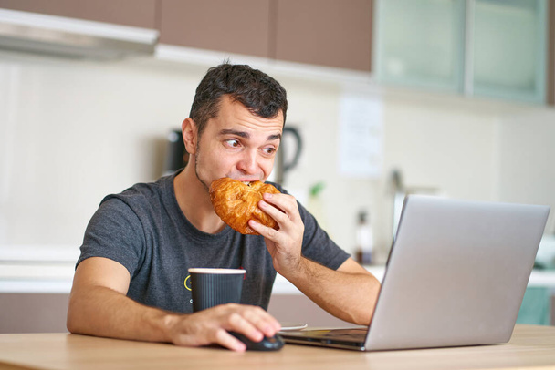 The guy has a croissant for breakfast and watches the morning news on his laptop. - Foto, Bild
