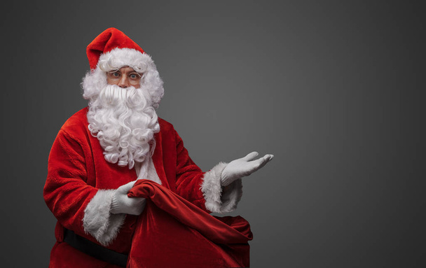 Aufnahme eines bärtigen Weihnachtsmannes mit riesiger Tasche und rotem Kostüm, der mit seiner Hand posiert. - Foto, Bild
