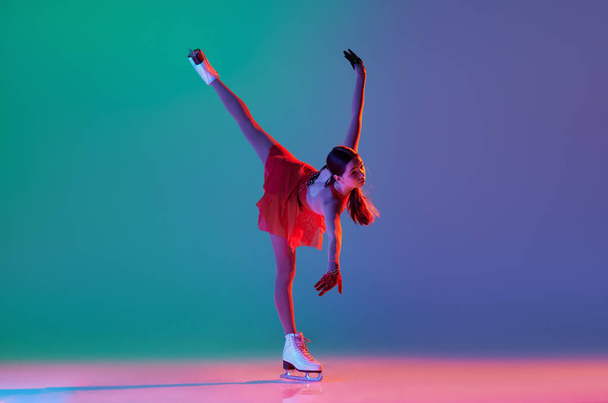 Young sportive girl, junior female figure skater in red stage costume skating isolated over gradient green-blue background in neon. Athlete in motion. Sport, beauty, winter sports. Copy space for ad - 写真・画像