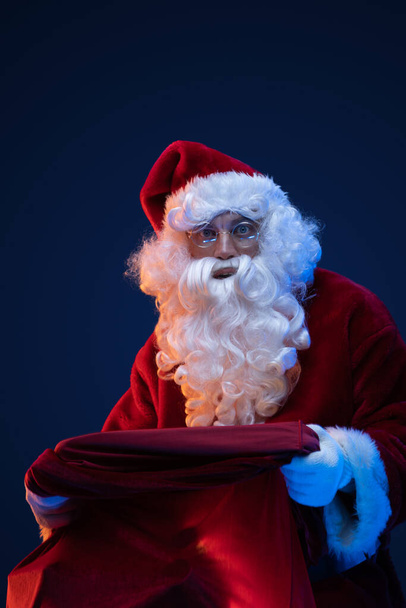 Portrait of bearded santa claus with red costume and bag against dark background. - Fotografie, Obrázek