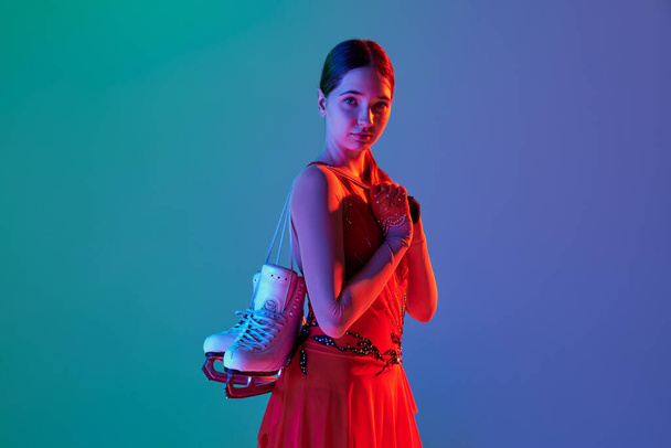 After workout. Young teen girl in stage costume dress standing with skates isolated over gradient green-blue background in neon light. Concept of skills, sport, hobbies, winter sports. Looks serious - 写真・画像