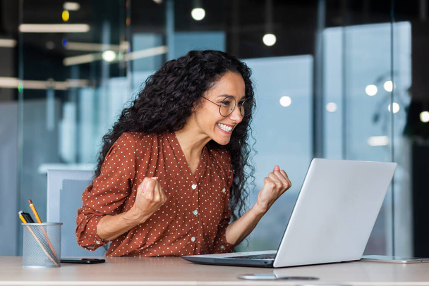 Hispanic business woman celebrating victory success, employee with curly hair inside office reading good news, using laptop at work inside office holding hand up and happy triumph gesture. - Photo, Image