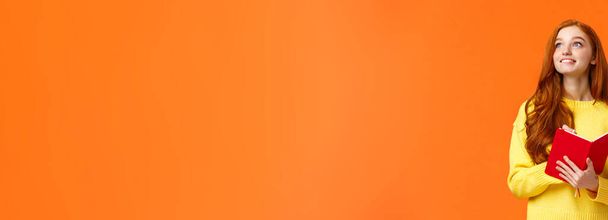 Dreamy, inspiradora linda pelirroja estudiante escribiendo tareas diarias, lista de tareas en rojo encantador planificador, mira esquina superior izquierda pensativo como sonriendo, inspirándose para escribir novela, pared naranja
. - Foto, imagen
