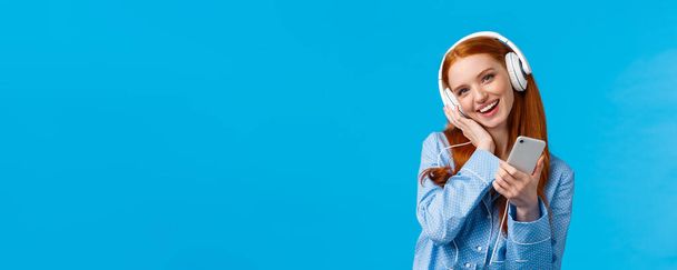 Lovely young dreamy girl fantasizing as listening favorite songs, voice of singer, tilt head and touching headphone as enjoying music earphones, holding smartphone and smiling, blue background. - Photo, Image