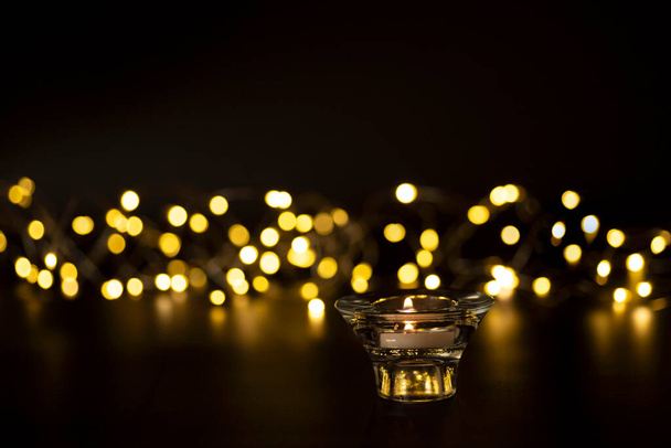 Burning candle in a glass holder, a glowing New Year's garland with blurry golden bokeh lights in the background. Concept of Christmas, romantic evening, wishes. Copy space. Nobody. - Photo, image