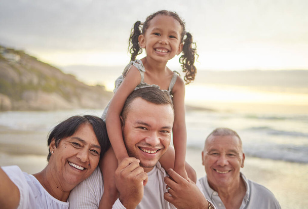 Happy mixed race family of four walking on the beach. A cute girl enjoying the sand and sea with their dad, grandfather and grandmother. Grandparents with their granddaughter and son outdoors. - Photo, Image
