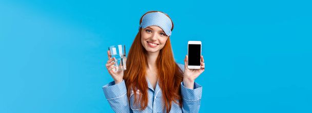 Waist-up πορτρέτο κομψό και glamour κοκκινομάλλα γυναίκα σε νυχτικά, μάσκα ύπνου, κρατώντας γυάλινο νερό και χαμογελώντας, κρατώντας smartphone, δείχνει το κινητό app στην οθόνη, μπλε φόντο. Αντιγραφή χώρου - Φωτογραφία, εικόνα