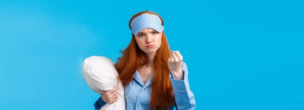Who disturbed my sleep. Mad and displeased, angry grumpy redhead woman threaten sibling being too loud in morning, shaking fist frowning, holding pillow, wearing pyjama and sleep mask. - Photo, Image