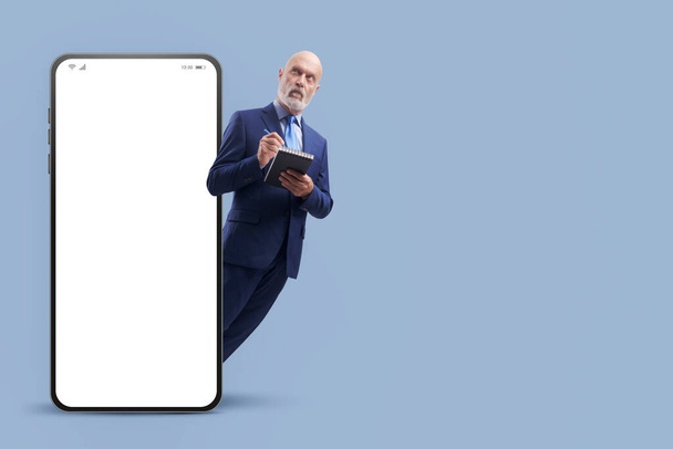 Corporate businessman writing down notes and big smartphone with blank screen, business and planning concept - Фото, зображення