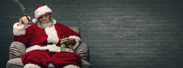 Bad Santa celebrating Christmas at home alone, he is smoking a cigar and drinking beer - Photo, image