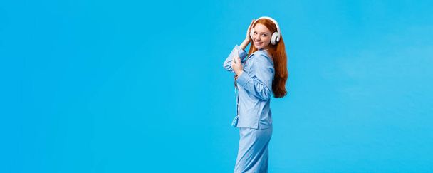 Vertical full-length portrait sassy and coquettish, flirty redhead woman in cute pyjama, dancing listen music in big white headphones, hold smartphone, using app to enjoy favorite playlist. - Foto, immagini