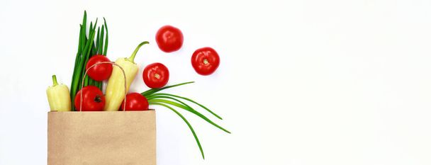 Gemüse wird aus einer Papiertüte auf weißem Hintergrund gegossen, Draufsicht. Das Konzept der gesunden Ernährung, des Einkaufs und der Bestellung von Lebensmitteln zu Hause mit Lieferung. Tomaten, grüne Zwiebeln und Paprika - Foto, Bild