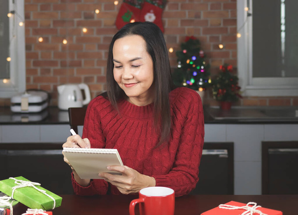 Portré ázsiai nő piros kötött pulóver ül asztalnál piros csésze kávé és ajándékdobozok a konyhában karácsonyi dekoráció, tollal írás névlista a notebook. - Fotó, kép