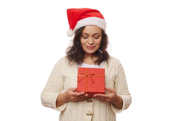 Красивая брюнетка в шляпе Санты и бежевом свитере, позирует с милой подарочной коробкой с счастливым подарком на Рождество в руках, изолированные на белом фоне. День бокса. Новый год. Копирование рекламного пространства - Фото, изображение