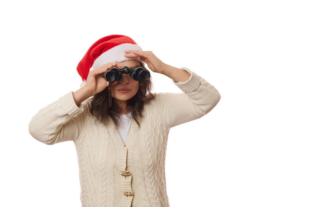 Donna attraente con cappello di Babbo Natale e maglione beige, guardando attraverso il binocolo, in previsione delle prossime vacanze invernali, isolato su sfondo bianco. Natale e Capodanno concetto. Copia spazio - Foto, immagini