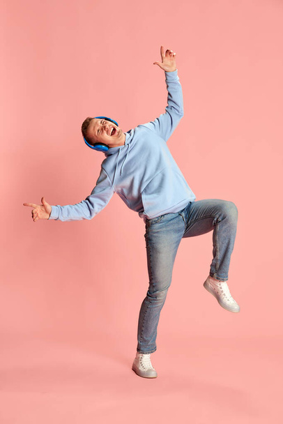 Portrait of young emotional man in hoodie and jeans listening to music in headphones and dancing isolated over pink background. Concept of youth, lifestyle, music, fashion, emotions, facial expression - Photo, Image