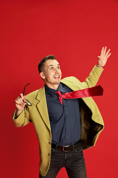 Portrait of young happy man in stylish jacket and glasses posing, showing excitement over red background. Win. Concept of emotions, business, occupation, hobby, lifestyle, fashion, facial expression - Foto, Imagem