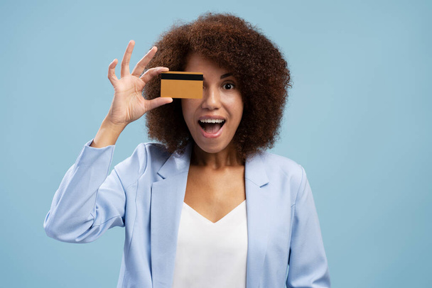 Giovane donna afroamericana stupito in possesso di carta di credito vicino al volto isolato su sfondo blu. Concetto shopping - Foto, immagini