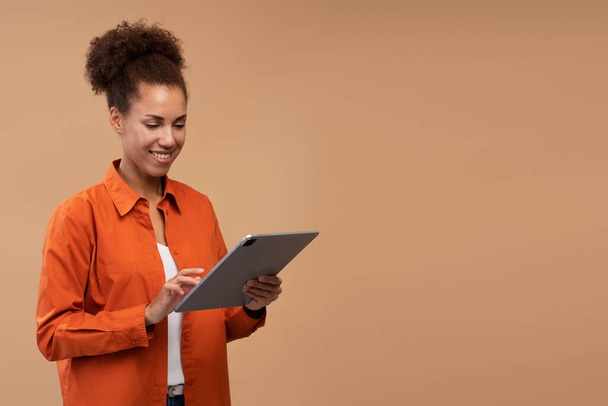 Uśmiechnięta Afroamerykanka używająca tabletu cyfrowego na beżowym tle, przestrzeń do kopiowania. Szczęśliwej udanej kobiety zakupy online. Koncepcja technologii  - Zdjęcie, obraz