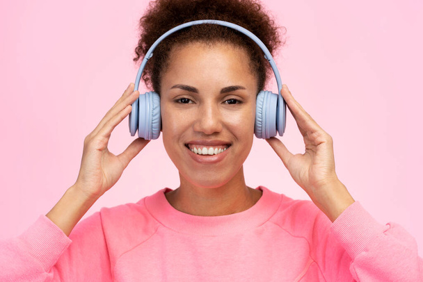 Sonriente mujer afroamericana con estilo escuchando música en auriculares inalámbricos mirando a la cámara aislada sobre fondo rosa. Concepto de tecnología moderna - Foto, Imagen