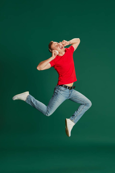 Portrait of young emotional man in casual clothes listening to music in headphones and jumping isolated over green background. Concept of youth, lifestyle, music, fashion, emotions, facial expression - Photo, Image