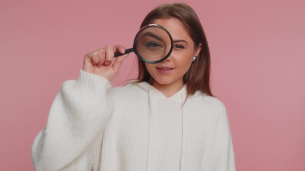 Investigator researcher scientist woman holding magnifying glass near face, looking into camera with big zoomed funny eyes, searching, analysing. Young adult girl isolated on pink studio background - Footage, Video
