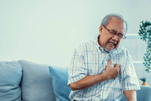 An agonizing senior man suffering from chest pain or heart attack alone in his living room. Serious health problem and feeling unwell concept for seniors. - Φωτογραφία, εικόνα