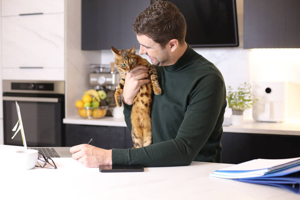 портрет красивого молодого человека с экзотическим котом, сидящим на кухне - Фото, изображение