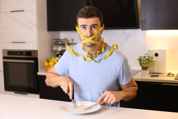 portrait of handsome young man with head tied in measuring tape at kitchen, diet concept - Photo, image