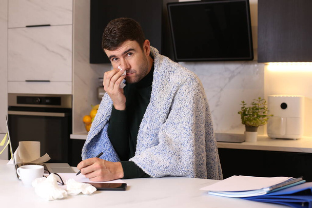 portrait of handsome young ill man with blanket working from home at kitchen - Photo, Image