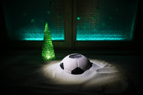 Παγκόσμιο Κύπελλο το χειμώνα έννοια. Ποδόσφαιρο (Ποδόσφαιρο) μπάλα σε χιονισμένο διακοσμημένο τραπέζι με μινιατούρες παιχνίδι. Πρωτοχρονιάτικο θέμα. Επιλεκτική εστίαση. Χαμηλό φως - Φωτογραφία, εικόνα