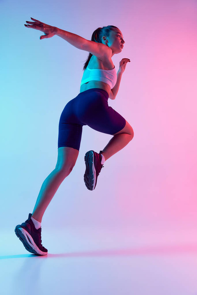 Bottom view of young sportive woman training, running isolated over gradient blue pink background in neon light. Concept of sport, strength, body care, fitness, wellbeing, health. Ad - Фото, изображение