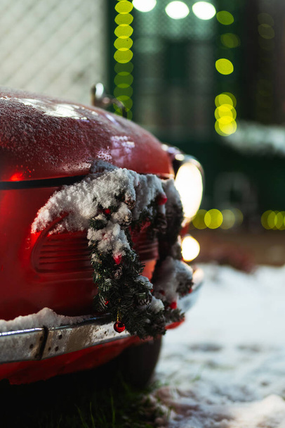 coche rojo con corona de Navidad y decoraciones. viejo coche rojo con decoraciones de año nuevo y nieve. Coche de Navidad para sesiones de fotos familiares en la calle - Foto, Imagen
