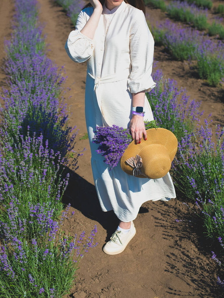 夏のフィールドに美しい紫色のラベンダーの花を歩くラベンダーの花束と麦藁帽子と白いリネンのドレスの女の子。暖かく、インスピレーションの概念。欲望と旅. - 写真・画像