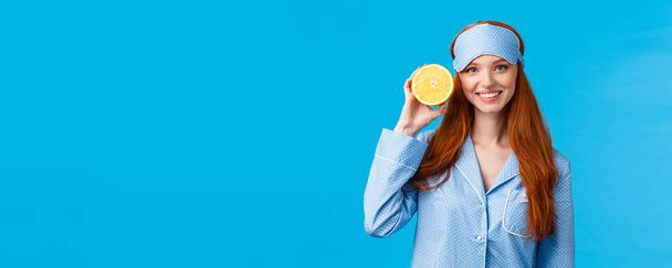 Start your morning right. Diet, healthy lifestyle and beauty concept. Cheerful glamour redhead woman in nightwear, holding slice orange near face, smiling, wearing sleep mask and pyjama. - Photo, Image