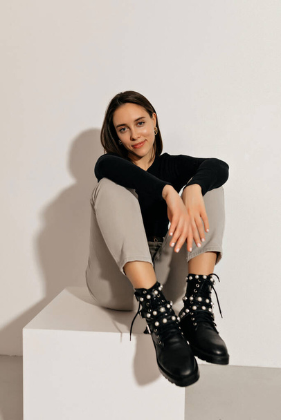 Full-lenght photo of stylish woman in black top, beige pants and black boots sitting on white chair over isolated background . - Zdjęcie, obraz