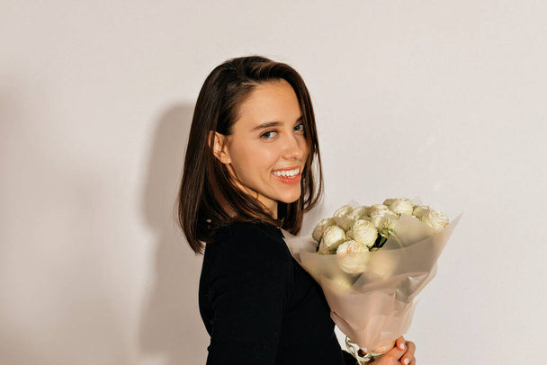 Sorridente ragazza attraente con i capelli scuri indossa camicia nera posa su sfondo isolato con fiori  - Foto, immagini